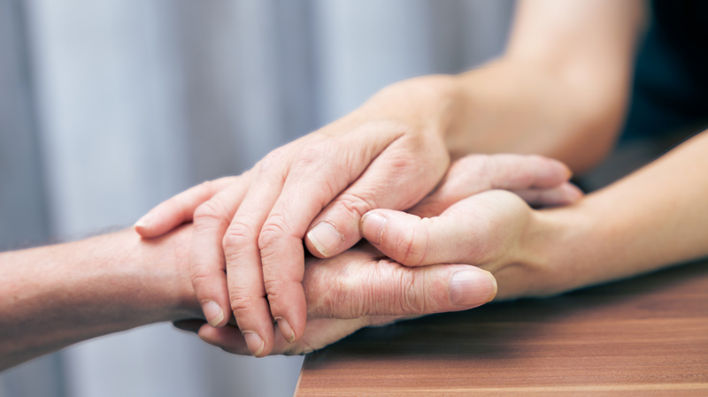 Nahaufnahme: Jüngere Hände halten fürsorglich die Hand einer älteren Person über einem Holztisch.