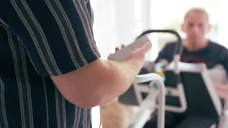 Person in gestreiftem Hemd reicht ein mobiles Gerät über einen Rollator an einen sitzenden, unscharfen Mann im Hintergrund.