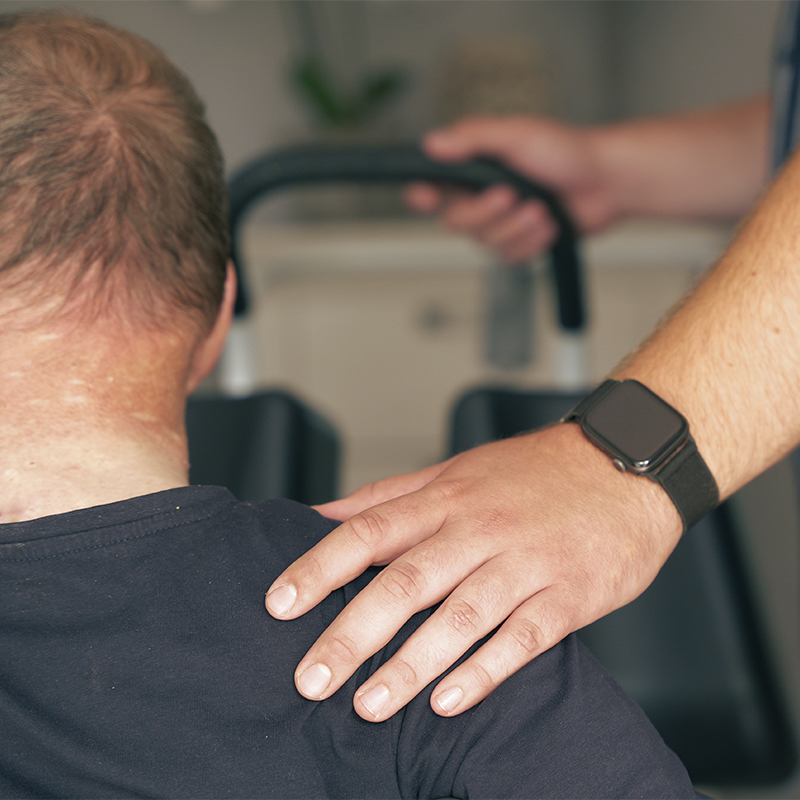 Hand mit Smartwatch liegt unterstützend auf der Schulter einer von hinten sichtbaren, dunkel gekleideten Person; im Hintergrund unscharfe Griffstangen eines Trainingsgeräts.
