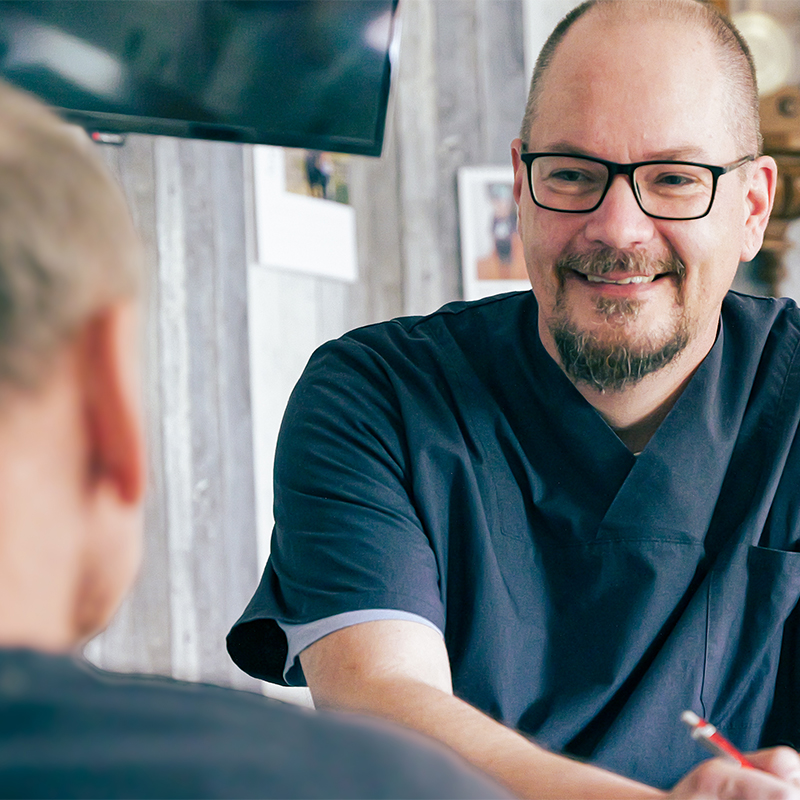 Lächelnder Mann mit Brille und kurzem Bart in dunkelblauer medizinischer Kleidung, der einem Patienten gegenüber sitzt und mit einem Stift in der Hand zuhört.