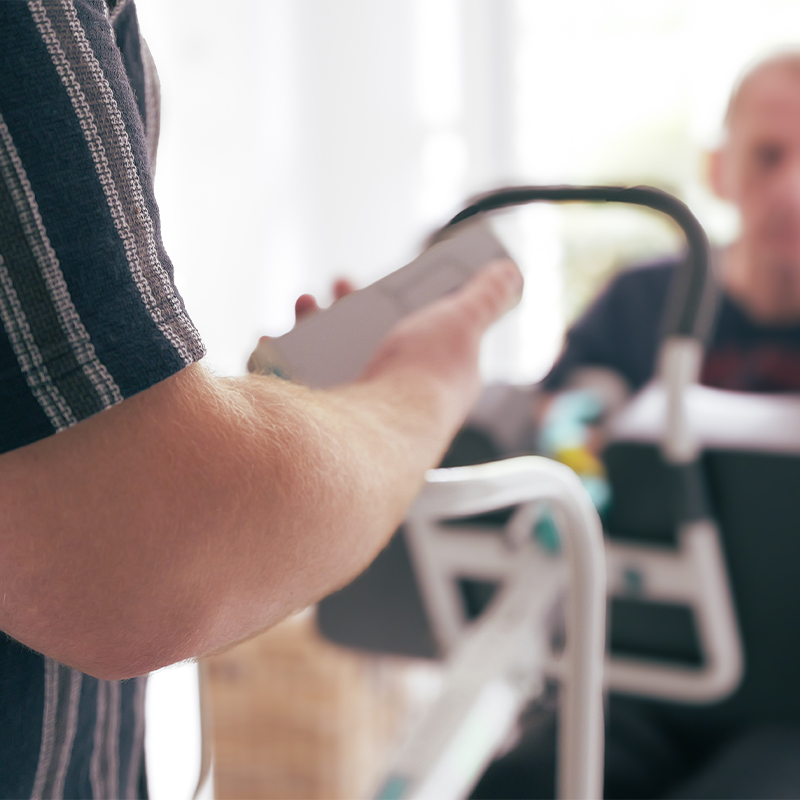 Nahaufnahme eines ausgestreckten Arms, der ein Smartphone über eine Gehhilfe hält; im Hintergrund unscharf eine sitzende Person.