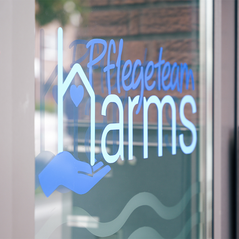 Blauer Aufkleber auf einer Glastür mit dem Schriftzug „Pflegeteam Harms“ und dem Symbol einer Hand, die ein Haus mit Herz hält, vor unscharfer Außenkulisse.