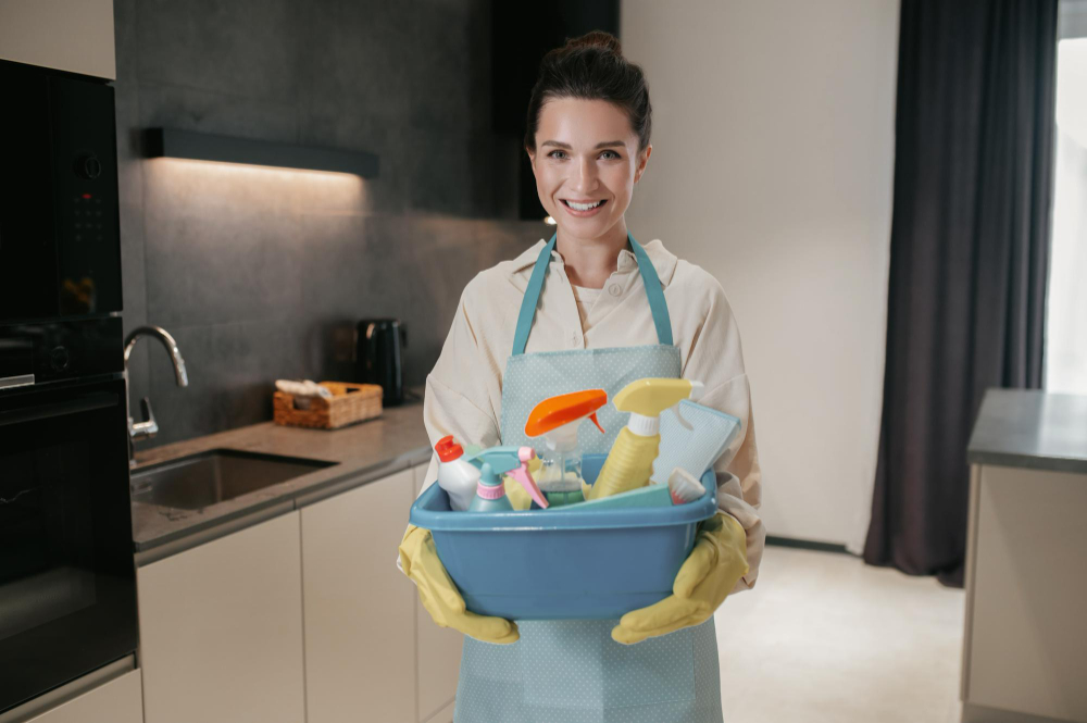 Lächelnde Frau in Schürze und gelben Gummihandschuhen hält in einer modernen Küche eine blaue Wanne mit verschiedenen Reinigungsflaschen und -utensilien.